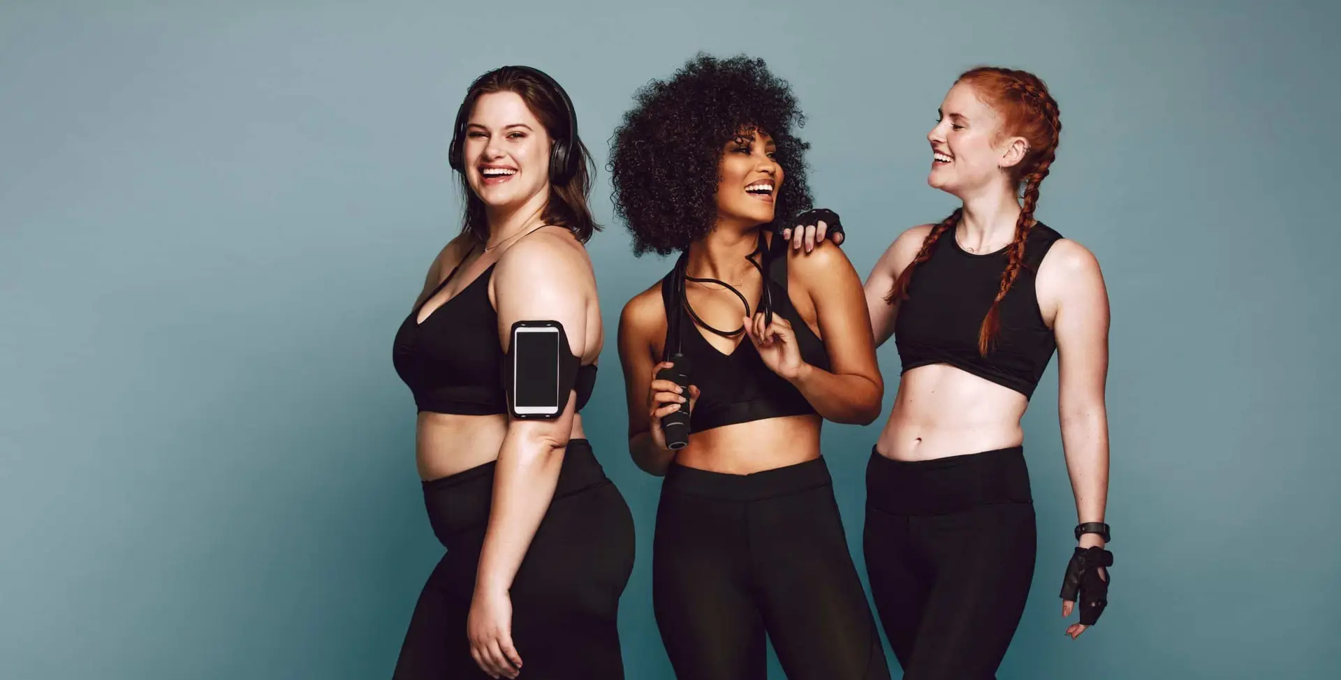 Trois femmes en tenue de sport noire rient ensemble ; l’une porte des écouteurs et son smartphone au bras, une autre tient une corde à sauter